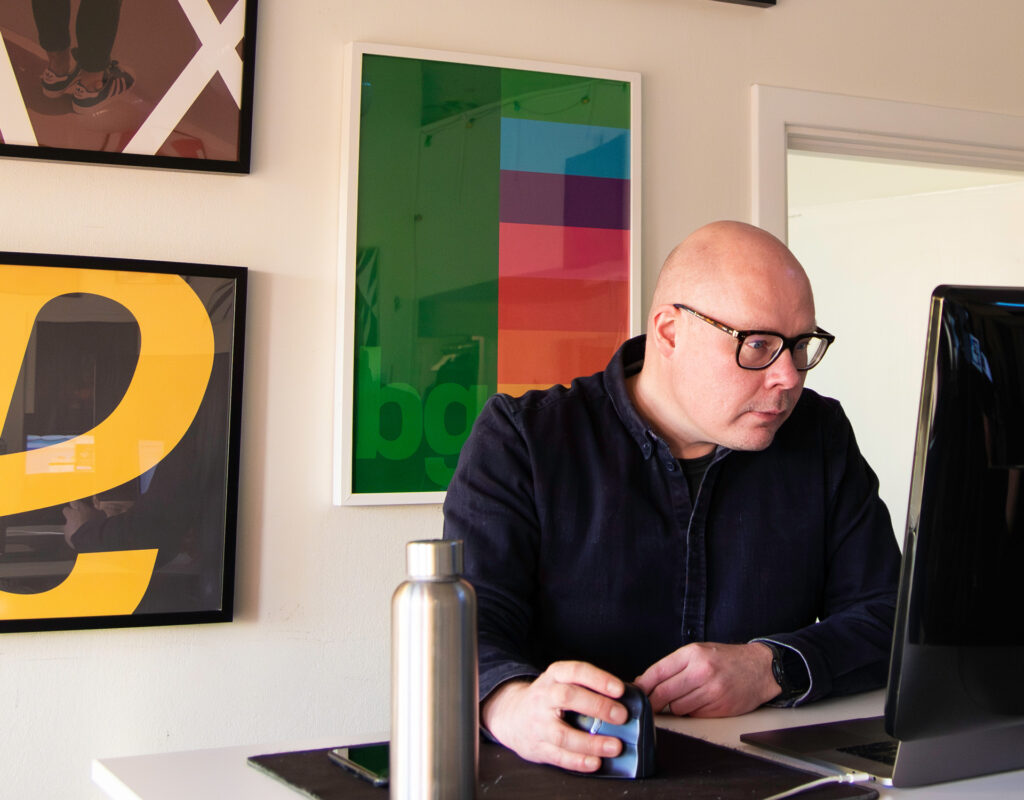 En person som jobbar vid en dator på en kreativ webbyrå. På väggen bakom honom syns färgglada tavlor. 