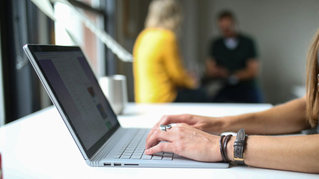 I förgrunden en kvinna som använder sin laptop och i bakgrunden sitter två personer och pratar med varandra